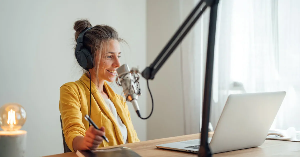 A woman in a yellow sweater sits at a desk with a laptop and headphones
